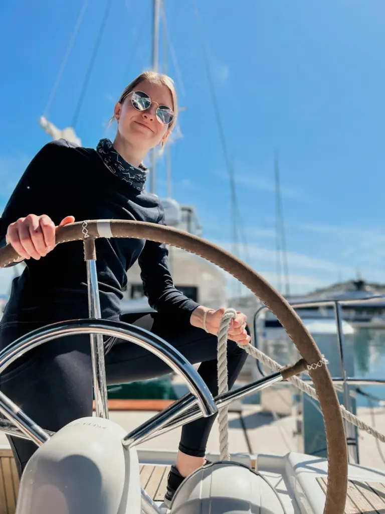 Frau mit schwarzer Kleidung und Sonnenbrille sitzt vor einem Steuerrad am Segelboot und blickt in die Kamera während sie Steuerrad und Tau in der Hand hält