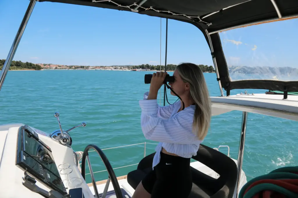 Frau steht am Segelboot am Meer und blickt mit Fernglas in die Ferne. Sie trägt eine schwarze Short und eine gestreifte weisse Bluse